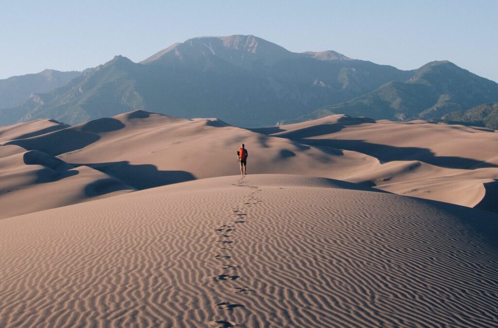 Man walking sand dunes national monument with mountains in the background